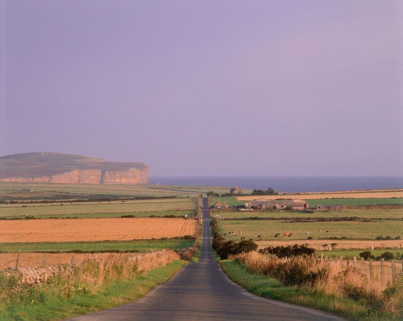 Looking Down A Straight Road Through Farmland With A View To Dunnet Head